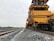 A large, yellow maintenance machine is positioned on the railway tracks, extending along the length of the tracks. The backdrop features some greenery under an overcast sky, and a person in an orange vest stands further down the tracks.