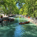 A turquoise-hulled electric canal boat gliding smoothly along a sunlit classic canal lined with greenery.