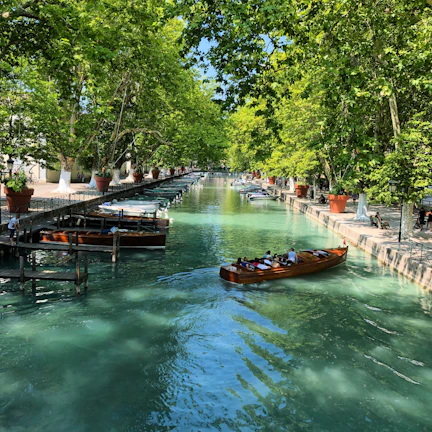A turquoise-hulled electric canal boat gliding smoothly along a sunlit classic canal lined with greenery.