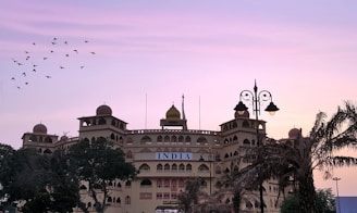 A large building with traditional architectural elements features multiple domes and arched windows. The facade displays the word 'INDIA' prominently. Palm trees and other vegetation partially obscure the view, while a flock of birds flies across a gradient sky transitioning from pink to purple. Street lamps add a decorative element to the scene.