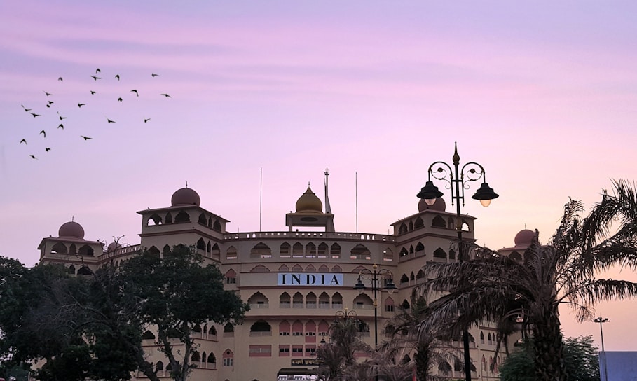 A large building with traditional architectural elements features multiple domes and arched windows. The facade displays the word 'INDIA' prominently. Palm trees and other vegetation partially obscure the view, while a flock of birds flies across a gradient sky transitioning from pink to purple. Street lamps add a decorative element to the scene.