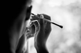 A monochrome photograph of a person holding a makeup brush close to their face. The person is captured from the side, focusing on their hand and the brush. The background appears blurred, suggesting a gentle and soft setting.