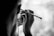 A monochrome photograph of a person holding a makeup brush close to their face. The person is captured from the side, focusing on their hand and the brush. The background appears blurred, suggesting a gentle and soft setting.
