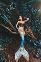A woman dressed as a mermaid with a brightly colored tail lies on a tree trunk in a dense, lush forest. The mermaid's scales display vibrant shades of blue and orange, contrasting with the earthy tones of the surrounding foliage. She is adorned with leaf-like coverings and has an elegant pose, emphasizing a mythical and fantasy theme.