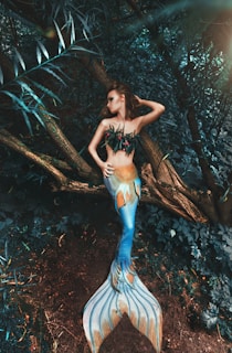 A woman dressed as a mermaid with a brightly colored tail lies on a tree trunk in a dense, lush forest. The mermaid's scales display vibrant shades of blue and orange, contrasting with the earthy tones of the surrounding foliage. She is adorned with leaf-like coverings and has an elegant pose, emphasizing a mythical and fantasy theme.