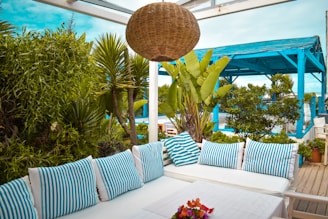 An outdoor seating area featuring a white sofa adorned with blue and white striped cushions. The space is surrounded by lush greenery, including potted plants and palm trees. A wicker light fixture hangs overhead, and a pergola structure with a bright blue frame is visible in the background. A small table with a floral arrangement in the center adds a touch of color.