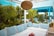 An outdoor seating area featuring a white sofa adorned with blue and white striped cushions. The space is surrounded by lush greenery, including potted plants and palm trees. A wicker light fixture hangs overhead, and a pergola structure with a bright blue frame is visible in the background. A small table with a floral arrangement in the center adds a touch of color.