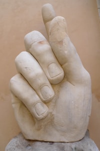 A large marble sculpture of a hand with detailed carvings of the fingers and fingernails. The sculpture has a smooth, weathered surface and is positioned against a neutral background.