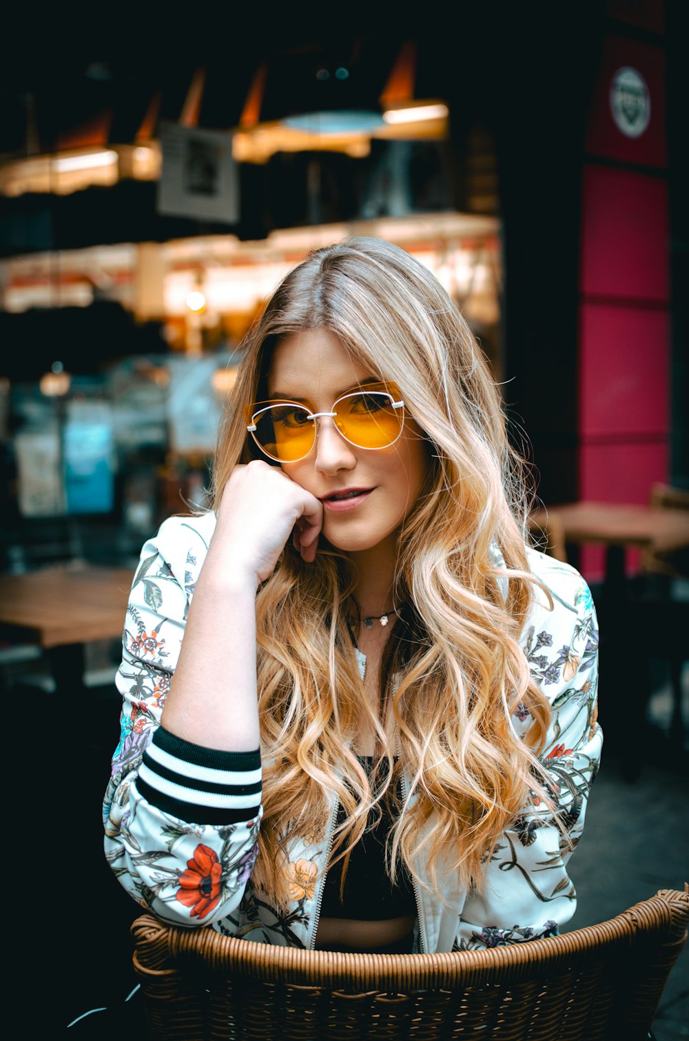 woman sitting on brown wicker chair holding her face wearing multicolored floral zip-up bomber jacket