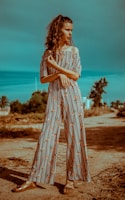 A woman stands confidently on a dirt path, wearing a long, striped jumpsuit with floral patterns. She has curly hair tied back and is looking to the side, with her arms folded in front of her. The background features green shrubbery and a clear blue sky, adding to a serene outdoor setting.