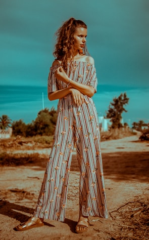 A woman stands confidently on a dirt path, wearing a long, striped jumpsuit with floral patterns. She has curly hair tied back and is looking to the side, with her arms folded in front of her. The background features green shrubbery and a clear blue sky, adding to a serene outdoor setting.