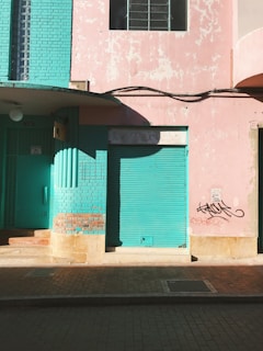 A facade featuring a combination of turquoise and pink painted brickwork. The wall has visible peeling and weathering effects. A closed turquoise shutter is centered, with graffiti on the pink section to the right. The pavement in front is made of red bricks, partially in shadow.