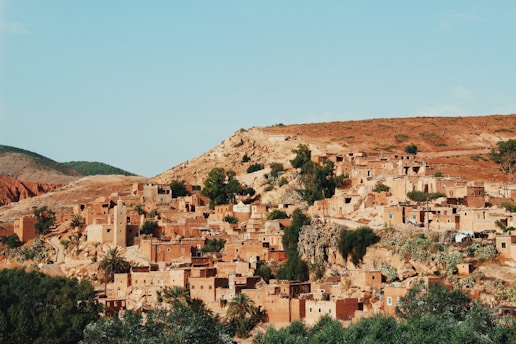 A cluster of traditional, earthy-tone buildings is nestled against a rocky hillside. The architecture blends seamlessly with the rugged landscape, which is dotted with sparse greenery and small trees. The warm tones of the buildings contrast with the clear blue sky, creating a picturesque scene. The village-like setting has a serene and timeless quality.