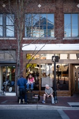 A sidewalk scene in front of a brick building with large windows. A man is shining a woman's shoes while an older man sits nearby on a chair with crutches. The area includes a streetlight and a tree with barren branches.