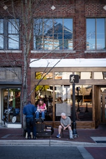 A sidewalk scene in front of a brick building with large windows. A man is shining a woman's shoes while an older man sits nearby on a chair with crutches. The area includes a streetlight and a tree with barren branches.