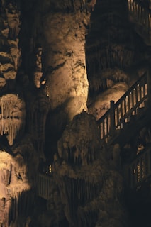 A captivating view inside a cave featuring intricate stalactite and stalagmite formations. A wooden staircase with railings is visible winding through the rocky interior, illuminated by soft, warm lighting that highlights the natural textures and structures.