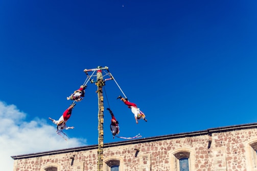A group of performers is engaged in a traditional dance, suspended from ropes and rotating around a tall pole against a clear blue sky. They are dressed in colorful traditional costumes, with a stone building visible at the bottom.