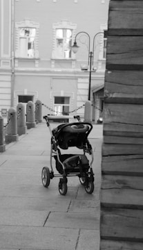 An empty baby stroller is placed on a paved walkway next to a series of stone posts connected by chains. The background features an old building with decorative windows, and there is a street lamp visible.