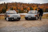 Two vintage cars are parked off-road near a lake. A person stands beside one car, examining it. The background features a scenic view of autumn foliage with trees in vibrant shades of orange and red. A couple of houses are visible across the lake under a cloudy sky.