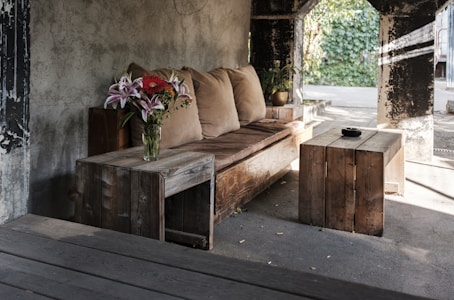 A cozy seating area with a rustic wooden sofa adorned with light brown cushions. A vase containing a vibrant bouquet of pink lilies and red flowers sits on a small wooden side table. Another wooden coffee table features a small black bowl. The setting is in a partially open space with a rough concrete wall and a view of lush greenery through large open windows.