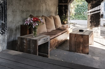 A cozy seating area with a rustic wooden sofa adorned with light brown cushions. A vase containing a vibrant bouquet of pink lilies and red flowers sits on a small wooden side table. Another wooden coffee table features a small black bowl. The setting is in a partially open space with a rough concrete wall and a view of lush greenery through large open windows.