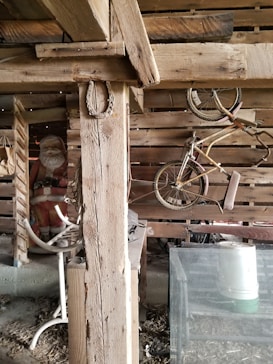Old wooden barn interior with rustic beams and wooden pallets. A vintage bicycle hangs from the wooden wall. A Santa Claus figure is partially visible in the background, adding a quirky touch. Visible are a white metal stool and some farm equipment.