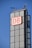 A tall, modern building structure features a prominent sign with the letters 'DB' in red on a white background. The building is composed of large glass panels with a grid-like appearance. The sky is clear and blue, indicating a bright day. Antennas and other technical installations are visible on top of the structure.