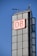 A tall, modern building structure features a prominent sign with the letters 'DB' in red on a white background. The building is composed of large glass panels with a grid-like appearance. The sky is clear and blue, indicating a bright day. Antennas and other technical installations are visible on top of the structure.