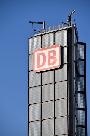 A tall, modern building structure features a prominent sign with the letters 'DB' in red on a white background. The building is composed of large glass panels with a grid-like appearance. The sky is clear and blue, indicating a bright day. Antennas and other technical installations are visible on top of the structure.