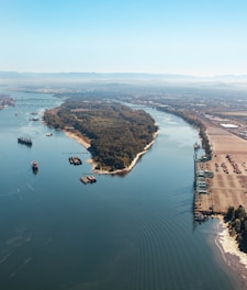 An aerial view of a large river with several islands covered in dense green foliage. Multiple cargo ships and barges are seen navigating the water. To the right, there is an industrial area with cranes and a parking lot. In the distance, mountains and a bridge spanning the river are visible under a clear blue sky.