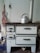 An old white metal stove with several drawers and a cooking surface, featuring a silver kettle placed on top. The stove has a pipe extending upwards, and there's a wall socket and hanging apron visible in the background.