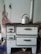 An old white metal stove with several drawers and a cooking surface, featuring a silver kettle placed on top. The stove has a pipe extending upwards, and there's a wall socket and hanging apron visible in the background.