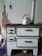 An old white metal stove with several drawers and a cooking surface, featuring a silver kettle placed on top. The stove has a pipe extending upwards, and there's a wall socket and hanging apron visible in the background.