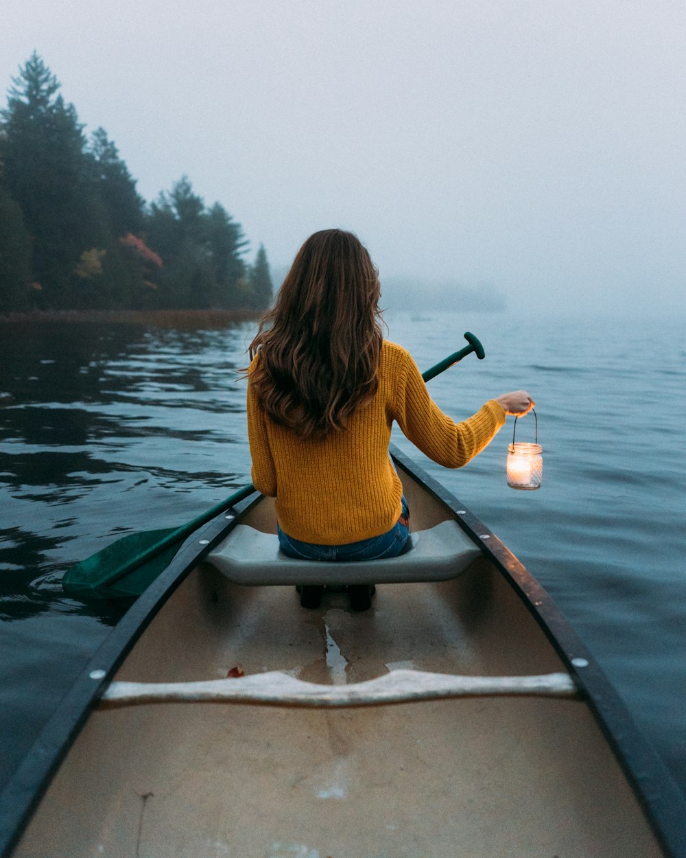Mujer sentada y sosteniendo lámpara en canoa de madera que ve cuerpo de agua tranquilo