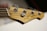 A close-up view of the headstock of a wooden electric bass guitar, showcasing tuning pegs and strings. The logo and some text are visible on the headstock, while the background is softly blurred.