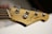 A close-up view of the headstock of a wooden electric bass guitar, showcasing tuning pegs and strings. The logo and some text are visible on the headstock, while the background is softly blurred.
