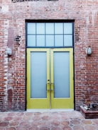 A pair of tall yellow doors with frosted glass panels set in a brick wall. Above the doors is a grid-patterned transom window. The brick wall appears weathered, with visible patches and two industrial-style wall lights flanking the doors. The ground is paved with hexagonal stone tiles.