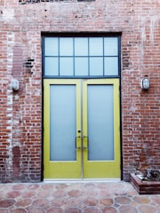 A pair of tall yellow doors with frosted glass panels set in a brick wall. Above the doors is a grid-patterned transom window. The brick wall appears weathered, with visible patches and two industrial-style wall lights flanking the doors. The ground is paved with hexagonal stone tiles.