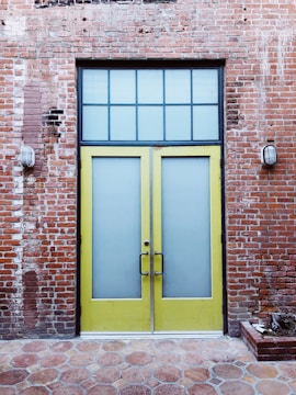 A pair of tall yellow doors with frosted glass panels set in a brick wall. Above the doors is a grid-patterned transom window. The brick wall appears weathered, with visible patches and two industrial-style wall lights flanking the doors. The ground is paved with hexagonal stone tiles.