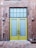 A pair of tall yellow doors with frosted glass panels set in a brick wall. Above the doors is a grid-patterned transom window. The brick wall appears weathered, with visible patches and two industrial-style wall lights flanking the doors. The ground is paved with hexagonal stone tiles.