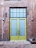 A pair of tall yellow doors with frosted glass panels set in a brick wall. Above the doors is a grid-patterned transom window. The brick wall appears weathered, with visible patches and two industrial-style wall lights flanking the doors. The ground is paved with hexagonal stone tiles.