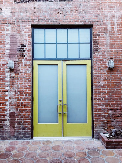 A pair of tall yellow doors with frosted glass panels set in a brick wall. Above the doors is a grid-patterned transom window. The brick wall appears weathered, with visible patches and two industrial-style wall lights flanking the doors. The ground is paved with hexagonal stone tiles.