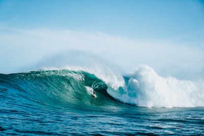A surfer skillfully rides a large, powerful turquoise wave. The wave is cresting with a white froth, and the ocean appears vast under a clear blue sky.