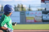 A baseball player wearing a green jersey and a blue helmet is captured in mid-action. The player is positioned in front of an out-of-focus background featuring a fence and several colorful advertisements. The player appears to be focused on the game, with hands positioned as if holding a bat or adjusting their gear.