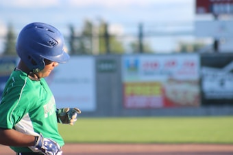 A baseball player wearing a green jersey and a blue helmet is captured in mid-action. The player is positioned in front of an out-of-focus background featuring a fence and several colorful advertisements. The player appears to be focused on the game, with hands positioned as if holding a bat or adjusting their gear.