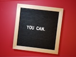 A wooden-framed letter board with a black background displays the motivational phrase 'YOU CAN.' in white letters, set against a vibrant red surface.