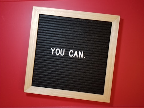 A wooden-framed letter board with a black background displays the motivational phrase 'YOU CAN.' in white letters, set against a vibrant red surface.