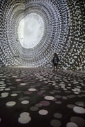 A person stands in a large, dark room surrounded by swirling patterns of white, circular lights projected on the walls and floor, creating an immersive tunnel-like visual experience.