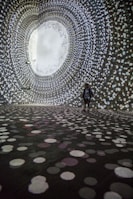 A person stands in a large, dark room surrounded by swirling patterns of white, circular lights projected on the walls and floor, creating an immersive tunnel-like visual experience.