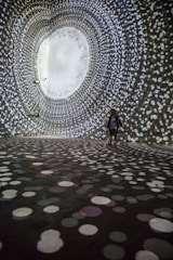 A person stands in a large, dark room surrounded by swirling patterns of white, circular lights projected on the walls and floor, creating an immersive tunnel-like visual experience.